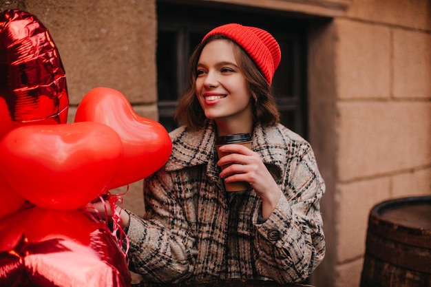 Mädchen mit leuchtend rotem Hut trinkt Kaffee Student wartet darauf, dass ein Freund ihm herzförmige Luftballons gibt