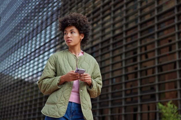 Mädchen mit lockigen Afro-Haaren nutzt Mobiltelefon für die Kommunikation in der Freizeit im Freien trägt Jacke und Jeans posiert im Freien genießt Erholungszeit beim Bummeln in der Stadtstraße