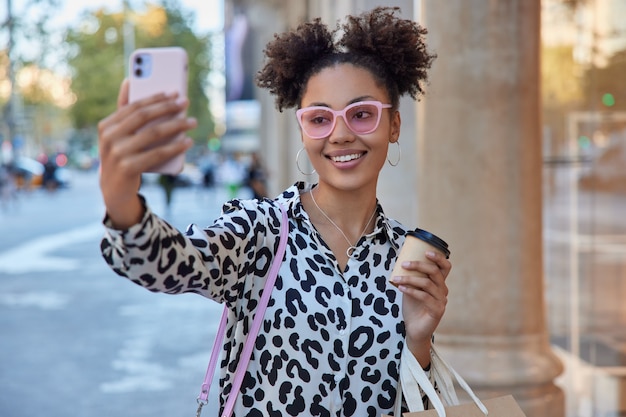 Mädchen mit lockigen Haaren spaziert draußen macht Selfie-Porträt auf Smartphone hält Kaffee zum Mitnehmen trägt rosa Sonnenbrille hat gute Laune nach dem Einkaufen macht Videoanrufe im Roaming