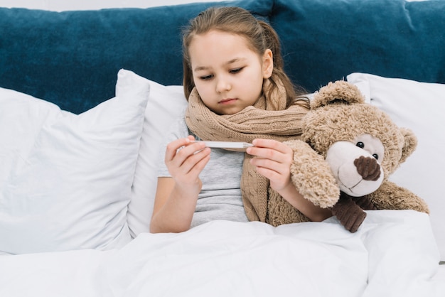 Mädchen mit Schal um ihren Hals, der mit dem Teddybären betrachtet Thermometer sitzt