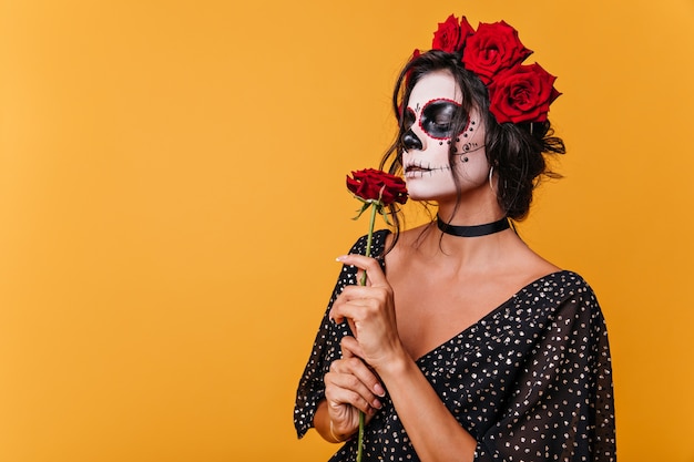 Mädchen mit Zombiemaske für Halloween riecht duftende Rose. Modell im schwarzen Kleid auf orange Wand