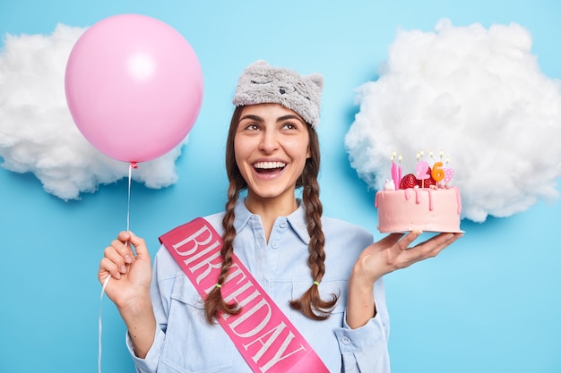 Mädchen mit zwei oben konzentrierten Zöpfen hat einen fröhlichen Ausdruck hält festlichen Kuchen und Heliumballon feiert den 26. Geburtstag wartet auf Freunde auf der Party nimmt Glückwünsche entgegen