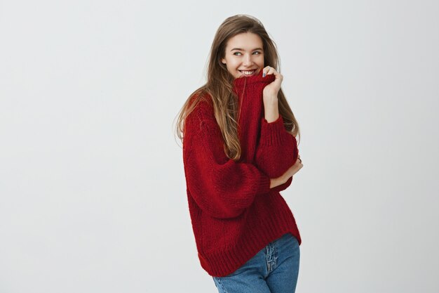 Mädchen mögen bequeme und lockere Kleidung. Studioaufnahme einer gut aussehenden charmanten Frau im roten Pullover, die den Kragen zieht, während sie halb gedreht steht, lächelt und mit flirtendem Ausdruck zur Seite schaut