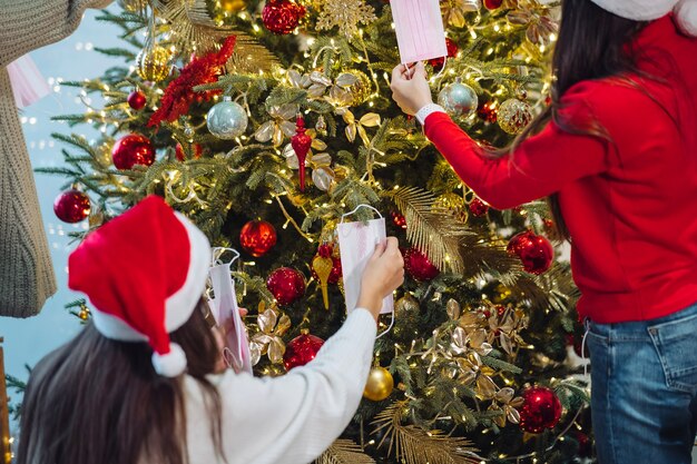 Mädchen schmücken den Weihnachtsbaum mit Schutzmasken. Weihnachten während des Coronavirus, Konzept