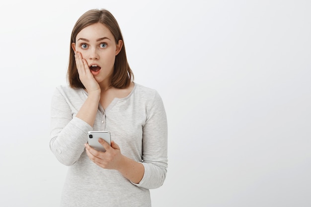 Mädchen schockiert zu sehen, peinliches Bild von Freund im Internet fallen Kiefer keuchen von überrascht halten Handfläche auf Wange starren erstaunt starren Smartphone über graue Wand