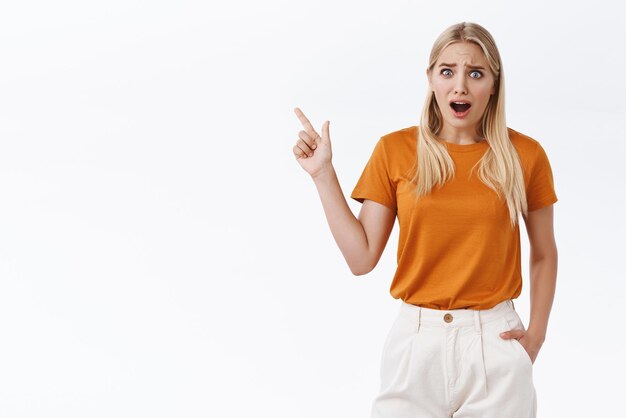 Mädchen schreckt vor ekelhafter Szene zurück Enttäuscht belästigte attraktive blonde Frau in orangefarbenem T-Shirt, die die Stirn runzelte und blinzelte, freakedout und frustriert auf den weißen Hintergrund der oberen linken Ecke