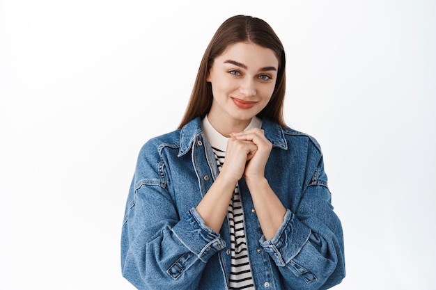 Mädchen sieht schüchtern und süß aus, macht ein albernes romantisches Gesicht, hält die Hände nah am Herzen und lächelt unschuldig in die Kamera, Engelsblick, auf weißem Hintergrund stehend