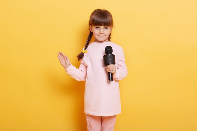 Mädchen singt mit Mikrofon und winkt Handfläche zur Kamera, lächelt, sieht süß und charmant aus, schaut in die Kamera, trägt Freizeitkleidung, Kind mit Zöpfen arrangiert Konzert für jemanden.