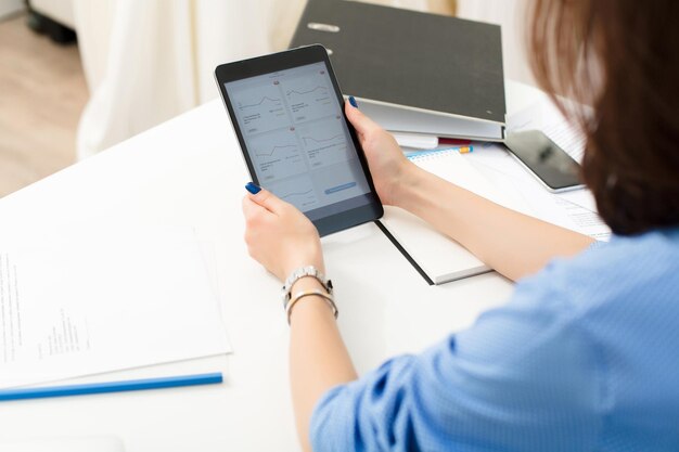 Mädchen sitzt am weißen Tisch im Büro. Sie hält Tablet und arbeitet mit Finanzdiagrammen.