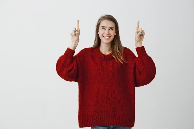 Mädchen träumt davon, im Wolkenkratzer zu leben. Verträumte attraktive weibliche Freundin in stilvollem, lockerem Pullover, die Zeigefinger hebt und nach oben zeigt, breit lächelt, interessiert und fasziniert ist