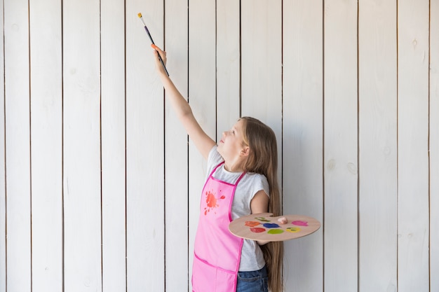 Mädchen, welches die hölzerne Palette versucht zu malen auf hölzerner Wand der weißen Planke hält