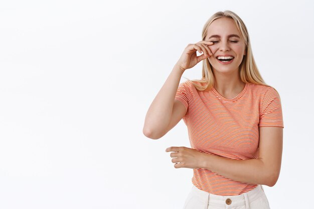 Mädchen wischt sich die Augenträne vom Lachen ab und genießt eine urkomische Komödie. Unbeschwerte, glückliche blonde Frau in gestreiftem T-Shirt, die Spaß hat, kichert, aufsteht, fröhlicher weißer Hintergrund steht