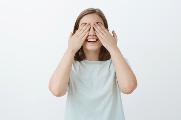 Mädchen zählt bis zehn bereit, Freunde zu suchen, während Verstecken spielen. Porträt der freudigen und emotionalen niedlichen Frau im hellblauen T-Shirt, das auf Überraschung mit geschlossenen Augen und Handflächen wartet, die lächeln