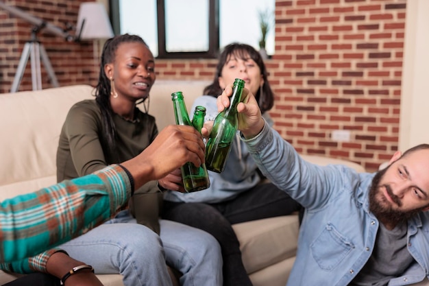 Männer und Frauen machen Jubelgesten und klirren mit Glasflaschen Bier und stoßen auf ein Wiedersehen der Freundschaft bei einem lustigen Treffen an. Fröhliche Menschen, die Toast mit alkoholischen Getränken machen, Freizeitaktivitäten.