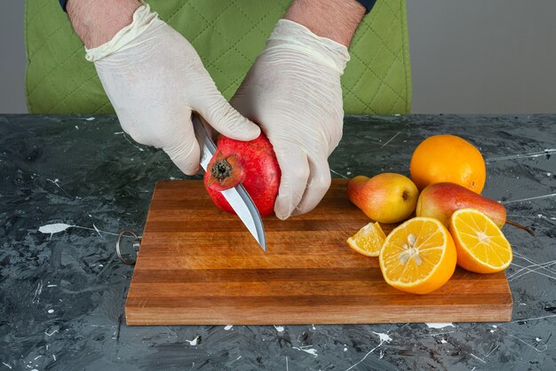 Männliche Hände, die roten Granatapfel auf Marmortisch schneiden.