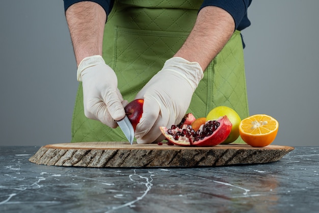 Männliche Hand in Handschuhen, die roten Granatapfel auf Marmortisch schneiden.