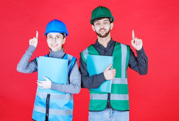 Männliche und weibliche ingenieure mit helmen, die blaue ordner halten und auf jemanden in der nähe zeigen.