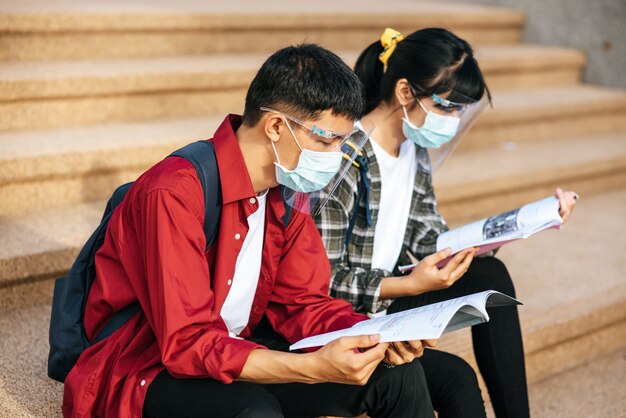 Männliche und weibliche Studenten, die Masken tragen, sitzen und lesen Bücher auf der Treppe