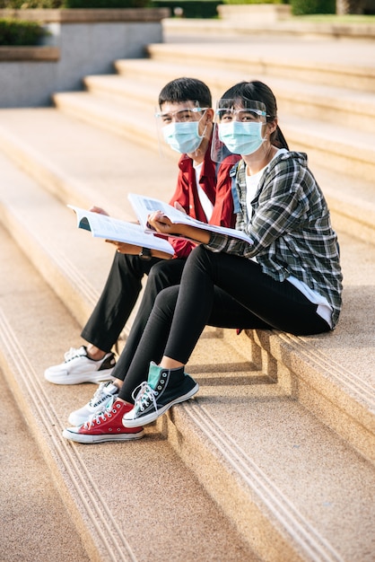 Männliche und weibliche Studenten, die Masken tragen, sitzen und lesen Bücher auf der Treppe