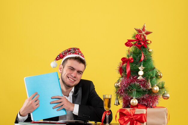 Männlicher Arbeiter der Vorderansicht, der Dokumente um kleinen Weihnachtsbaum und Geschenke auf Gelb hält