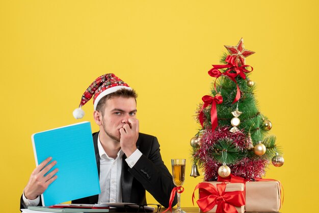 Männlicher Arbeiter der Vorderansicht, der Dokumente um kleinen Weihnachtsbaum und Geschenke auf Gelb hält