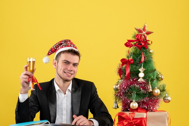 Männlicher Arbeiter der Vorderansicht, der Weihnachten mit Champagner um kleinen Weihnachtsbaum feiert und auf Gelb präsentiert