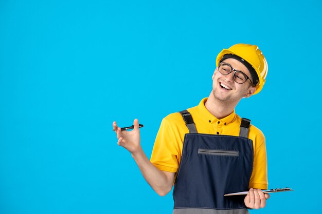 Männlicher Arbeiter der Vorderansicht in der Uniform und im Helm, die Notizen machen und auf Blau lachen