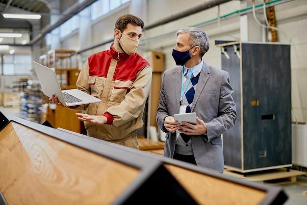 Männlicher Arbeiter, der während der Coronavirus-Pandemie mit dem Firmenleiter in der Holzfabrik kommuniziert, benutzt Laptop