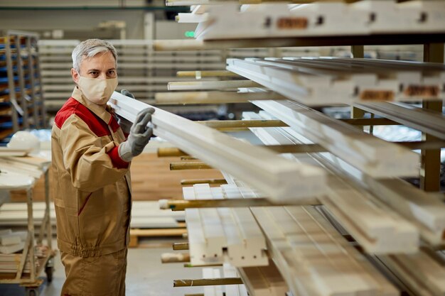 Männlicher Arbeiter mit Gesichtsmaske, der Bretter auf einem Gestell in der Tischlerei stapelt