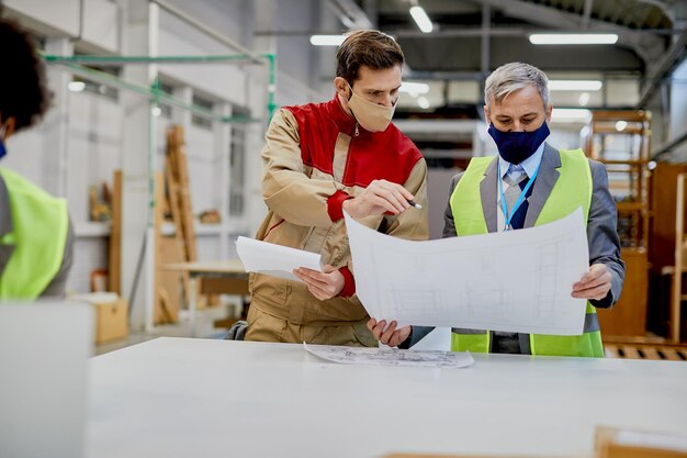 Männlicher Arbeiter und Geschäftsmann mit schützenden Gesichtsmasken, der Blaupausen in der Holzbearbeitungsfabrik analysiert