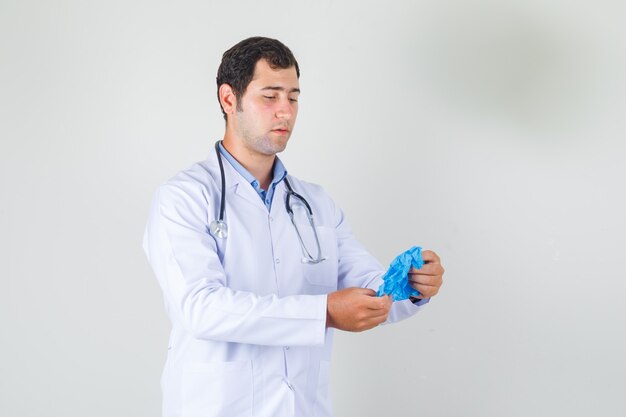 Männlicher Arzt, der blaue medizinische Handschuhe im weißen Kittel hält und vorsichtig schaut. Vorderansicht.