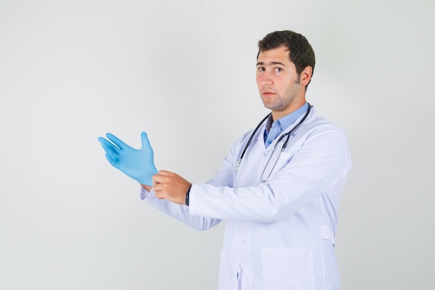Männlicher Arzt, der blaue medizinische Handschuhe im weißen Kittel trägt und ernst schaut. Vorderansicht.