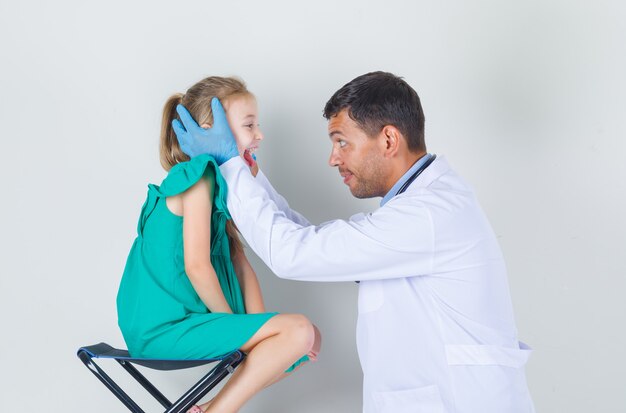 Männlicher Arzt, der den Hals des Kindes in der weißen Uniform untersucht und fröhlich aussieht. Vorderansicht.
