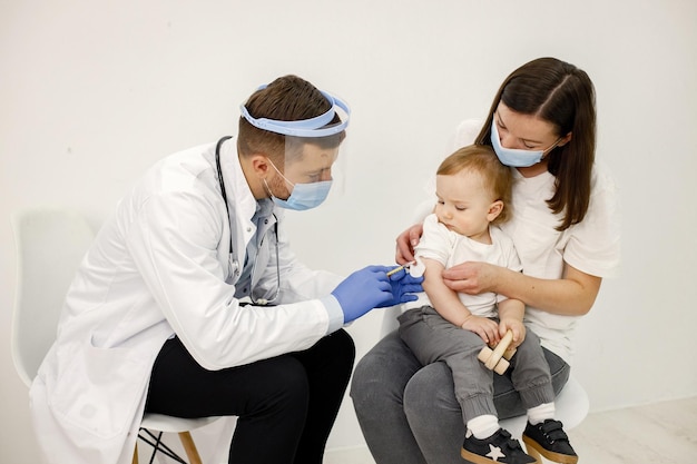 Männlicher Arzt, der einem kleinen Jungen, der auf dem Schoß der Mutter sitzt, eine Impfung vornimmt