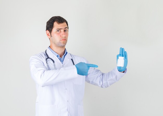 Männlicher Arzt, der Finger auf Flasche der Pillen im weißen Kittel, Handschuhe zeigt und streng schaut