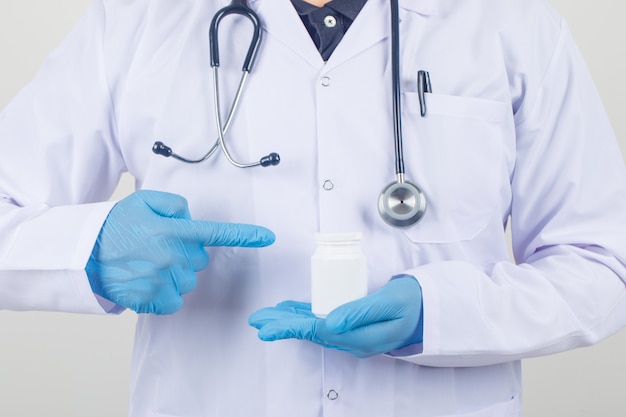 Männlicher Arzt, der Finger auf Pillenflasche im weißen Kittel und in den Handschuhen zeigt und vorsichtig schaut