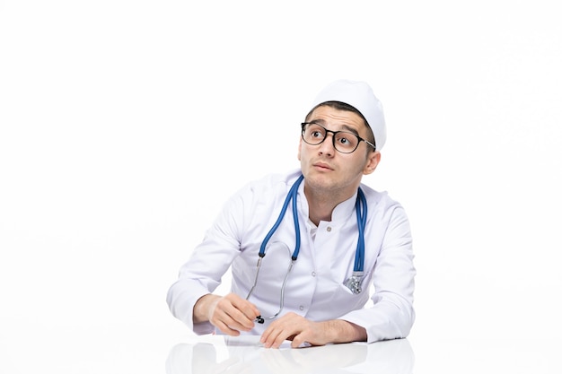 Männlicher Arzt der Vorderansicht im medizinischen Anzug, der hinter Schreibtisch sitzt
