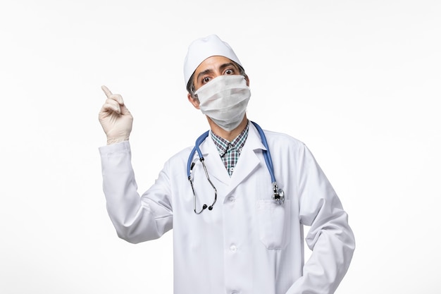 Männlicher Arzt der Vorderansicht im medizinischen Anzug und in der Maske wegen Coronavirus auf weißem Schreibtisch