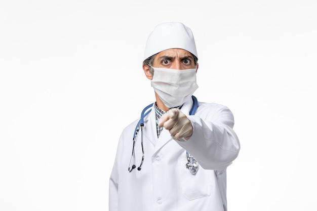 Männlicher Arzt der Vorderansicht im weißen medizinischen Anzug, der Maske wegen Covid auf hellweißem Schreibtisch trägt
