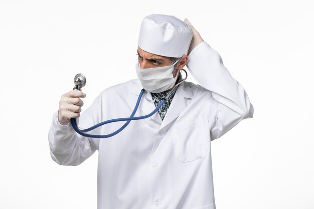 Männlicher Arzt der Vorderansicht im weißen medizinischen Anzug, der Maske wegen des Coronavirus hält, der Stethoskop auf weißer Oberfläche hält