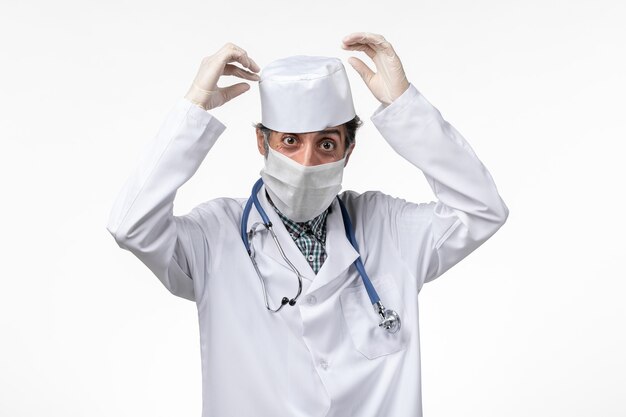 Männlicher Arzt der Vorderansicht im weißen medizinischen Anzug mit Maske wegen Covid auf weißem Schreibtisch