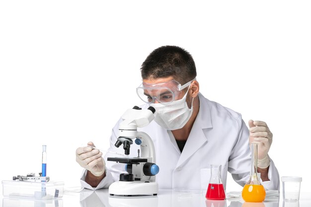 Männlicher Arzt der Vorderansicht im weißen medizinischen Anzug mit Maske wegen Covid unter Verwendung des Mikroskops auf weißem Raum