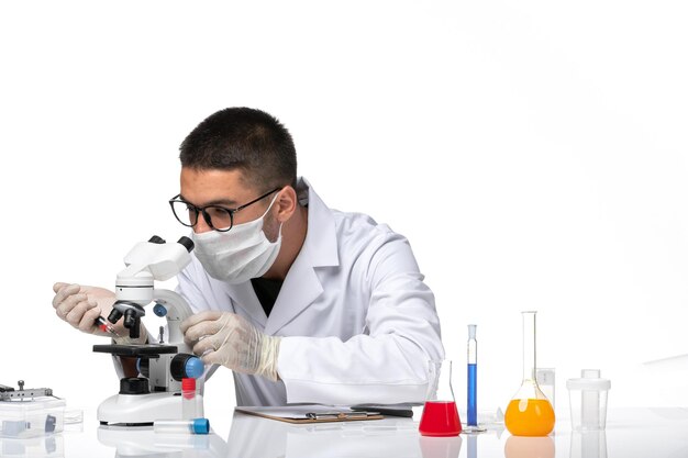 Männlicher Arzt der Vorderansicht im weißen medizinischen Anzug mit Maske wegen der koviden Arbeit mit dem Mikroskop auf dem weißen Raum