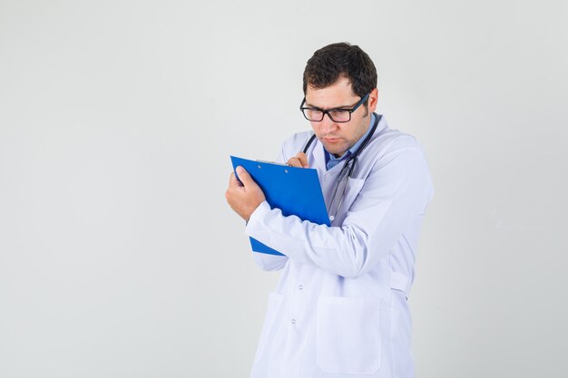 Männlicher Arzt im weißen Kittel, Brille, die Notizen auf Zwischenablage macht und ernst schaut