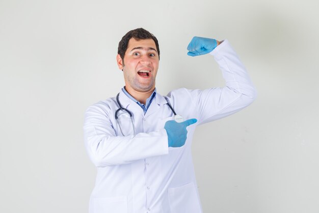Männlicher Arzt im weißen Kittel, Handschuhe, die Muskeln mit Finger zeigen und stark schauen