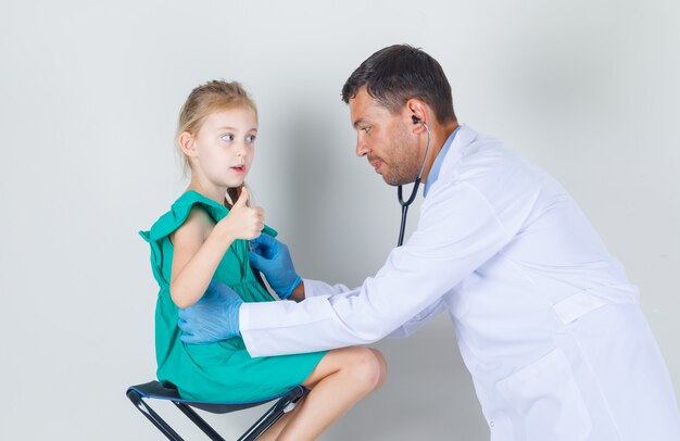 Männlicher Arzt in der weißen Uniform, die Herzschlag hört, während Kind Daumen zeigt