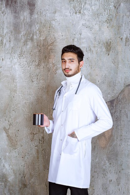 Männlicher Arzt mit Stethoskop, das eine Tasse Kaffee hält.