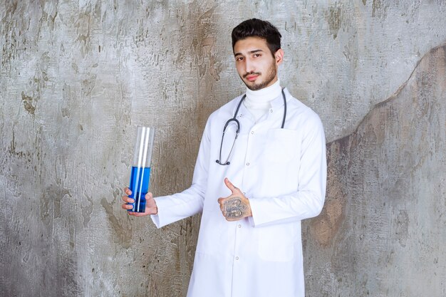 Männlicher Arzt mit Stethoskop, der einen chemischen Kolben mit blauer Flüssigkeit nach innen hält und erfolgreiches Handzeichen zeigt.