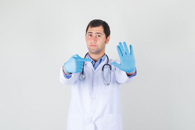 Männlicher Arzt zeigt Finger auf seine Handfläche im weißen Kittel, Handschuhe und sieht ernst aus