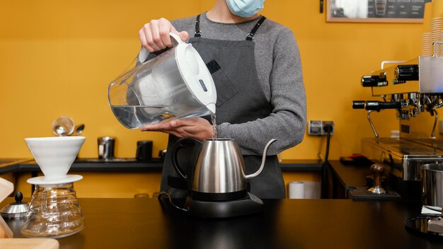 Männlicher Barista mit medizinischer Maske, die Wasser in den Kessel gießt